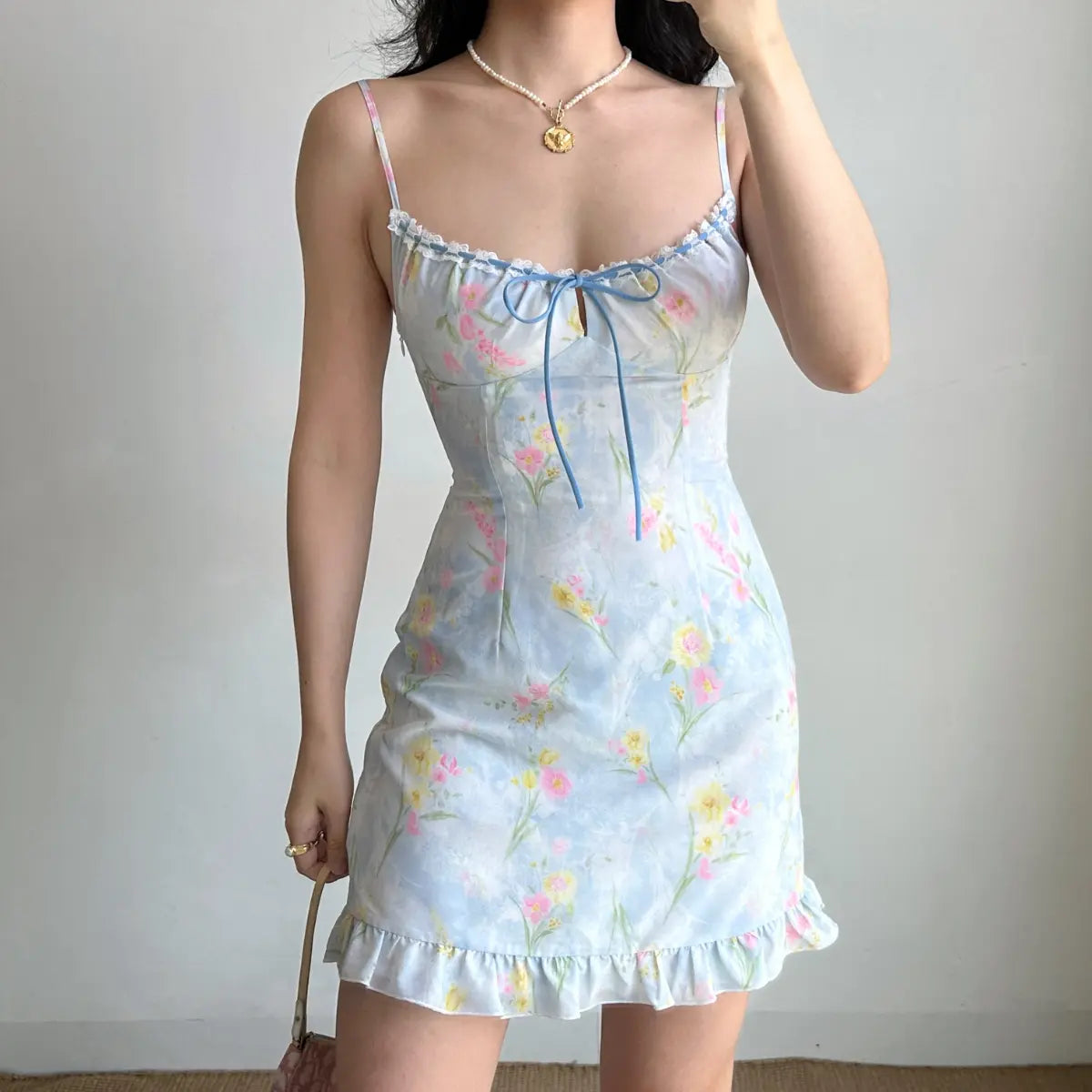  Woman wearing a light blue watercolor floral mini dress, accessorized with a pearl necklace, showing the dress’s fitted bodice and ruffled hem.
