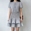 Model wearing a light gray plaid dress with a smocked waist, puff sleeves, and a tiered skirt, carrying a brown handbag.