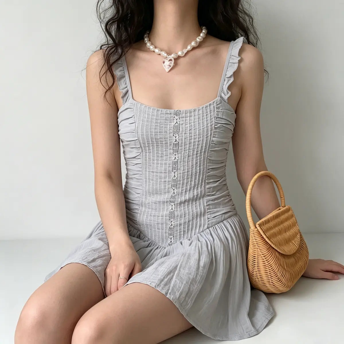 Seated view of a model wearing a light gray ruched mini dress, with a rattan bag beside her.