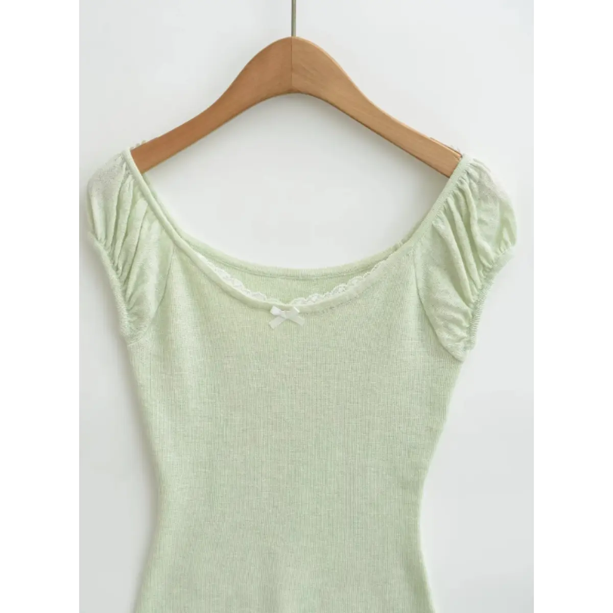 Front view of a light green ribbed off-the-shoulder top hanging on a wooden hanger, showing the wide scoop neckline, lace trim, and small white bow.