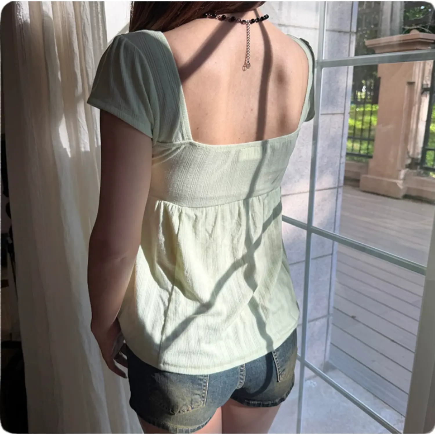 Back view of a woman wearing a light green square-neck puff sleeve top, standing by a sunlit window