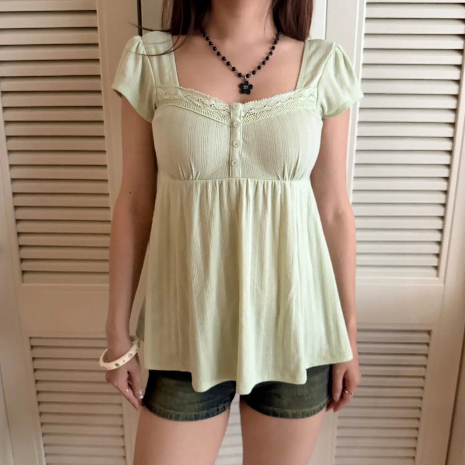 Full-body shot of a woman in a light green square-neck puff sleeve top with lace trim and buttons, paired with denim shorts