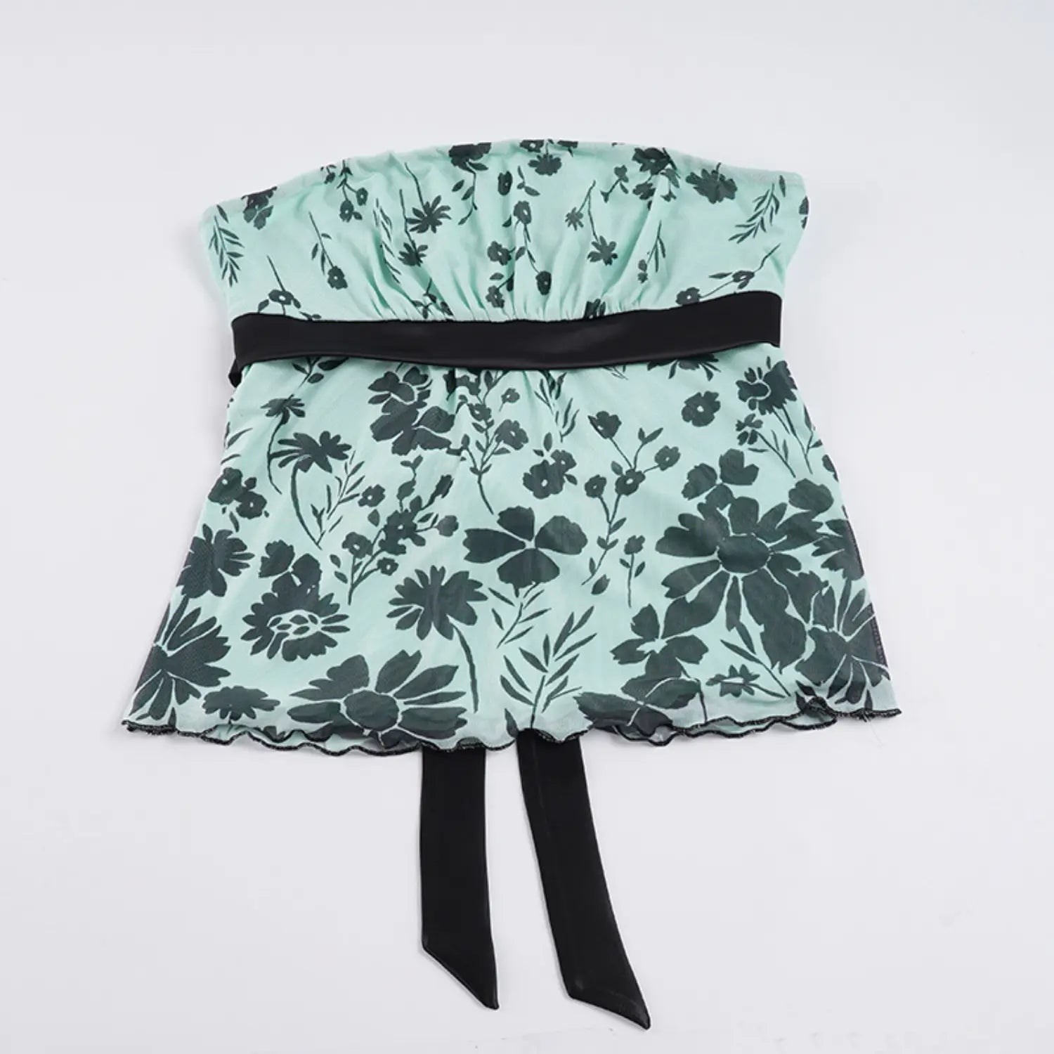 Full flat lay studio shot of strapless mesh tube top on plain white background, elastic gathered bust panel, solid black satin waist ribbon with two long hanging pointed ties, scalloped wavy mesh hem, scattered black floral print