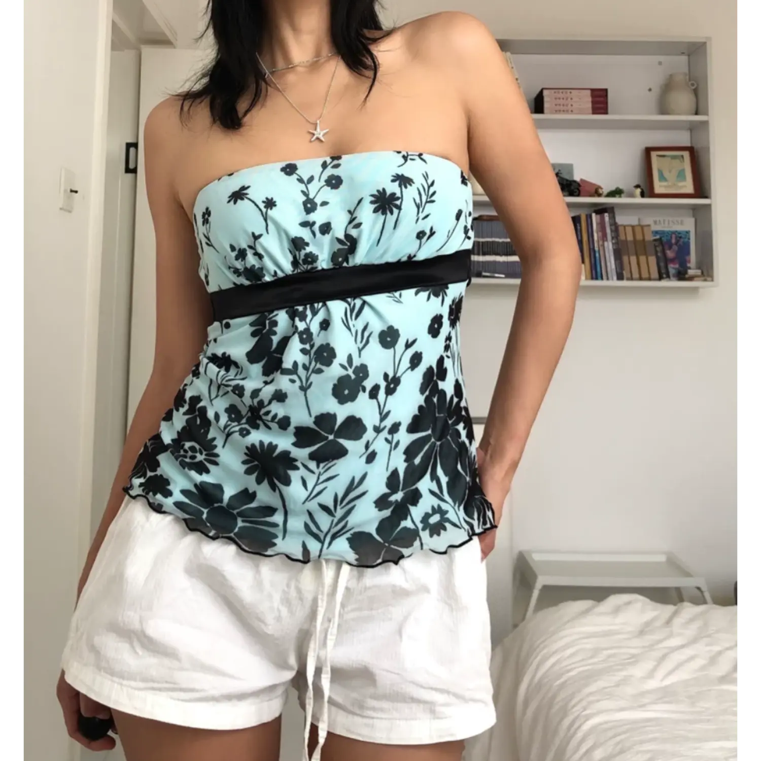 Casual indoor bedroom lifestyle shot of sheer mint mesh strapless top with black floral print, model stretching one arm out naturally, layered silver star pendant necklace, matching loose white casual shorts, soft natural room lighting