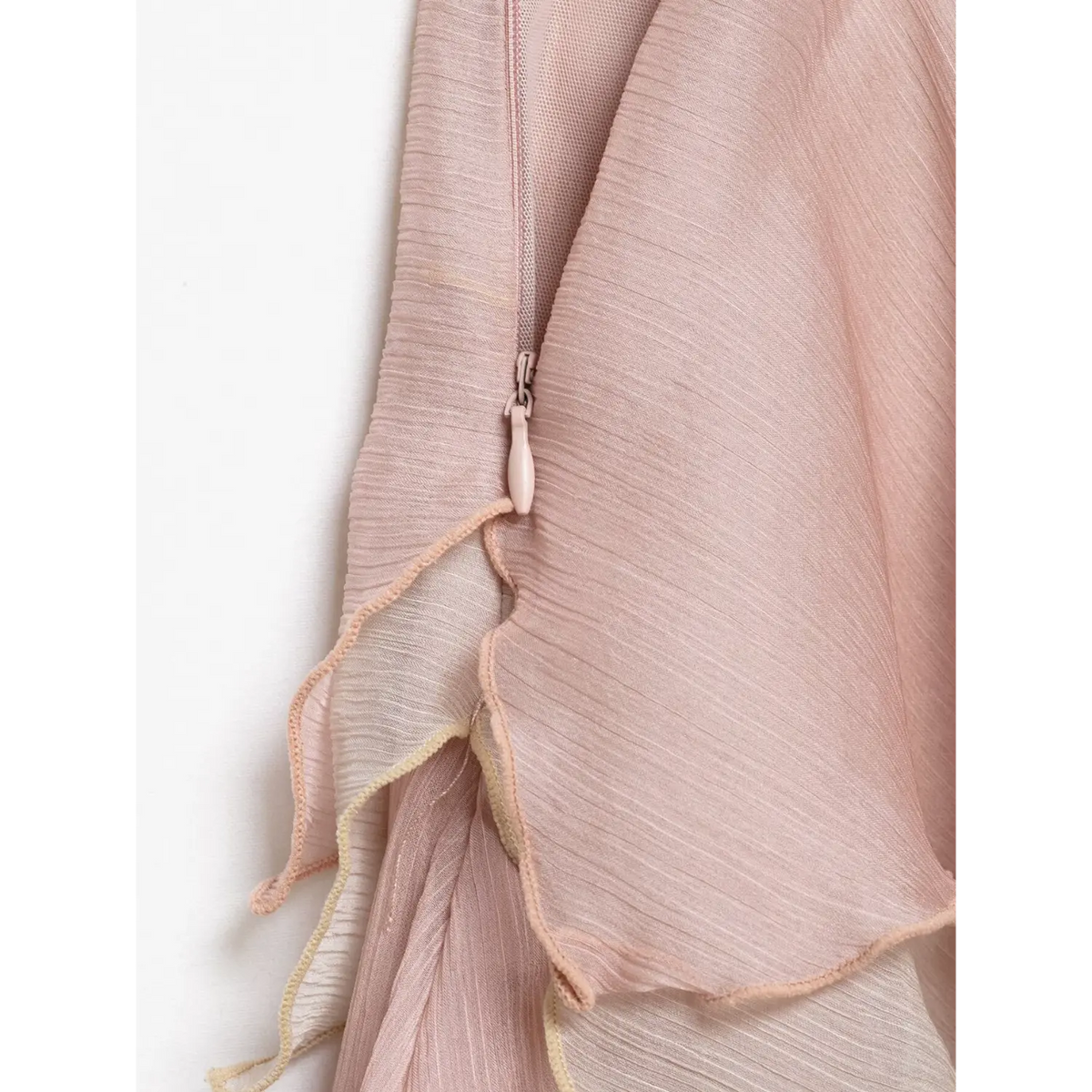Close-up of invisible side zipper on light pink chiffon skirt, matching pink zipper pull beside layered ruffle trim, white backdrop