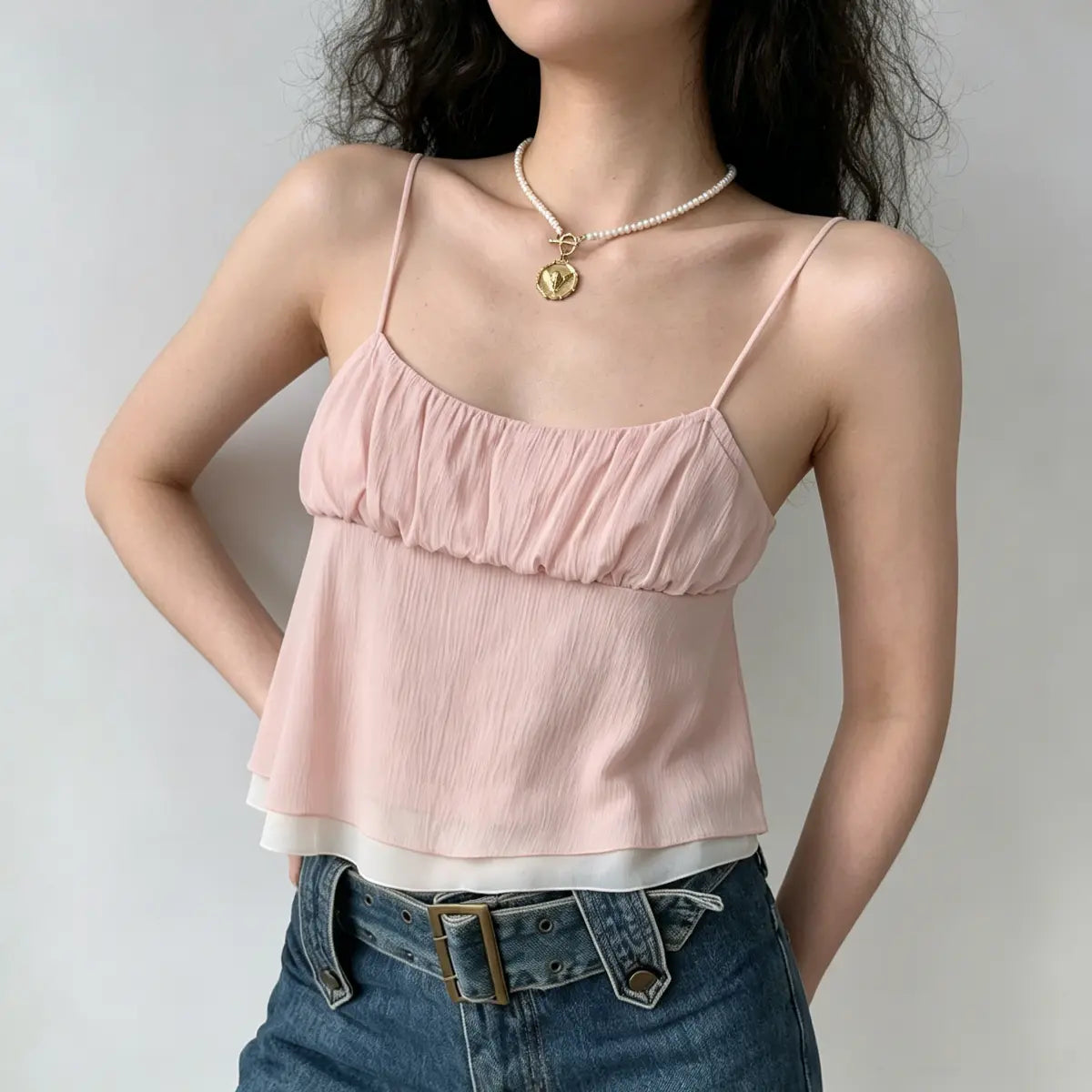Model wearing a light pink double-layer ruffled cami top with thin spaghetti straps, featuring a gathered bust and flowy silhouette, paired with jeans.