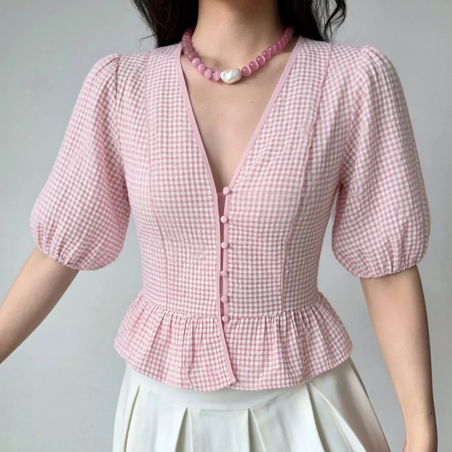 Front lifestyle shot on model, v-neck puff short sleeve gingham blouse, front button placket, peplum ruffle hem, matched with pink bead necklace and white pleated skirt, sweet casual summer outfit
