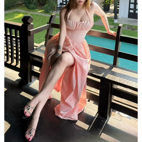 Full view of a woman in a light pink halter maxi dress by a pool, displaying the halter straps, corset waist, and flowing skirt.