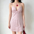 Model wearing a light pink spaghetti-strap mini dress with a bustier-style bodice, center knot detail, and vertical pleats, styled with a red pendant necklace and red handbag, front view.
