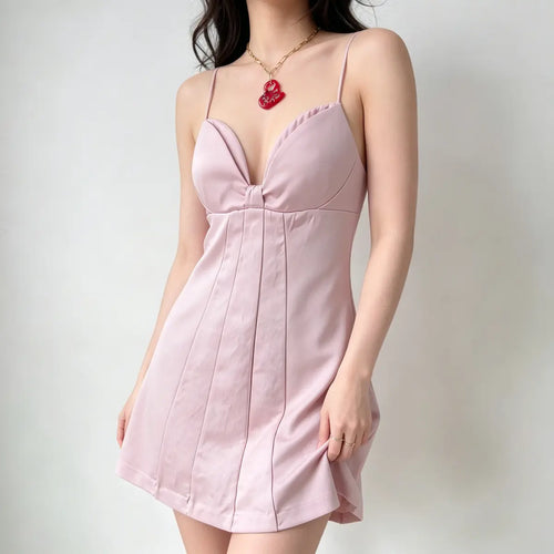 Model wearing a light pink spaghetti-strap mini dress with a bustier-style bodice, center knot detail, and vertical pleats, styled with a red pendant necklace and red handbag, side view.