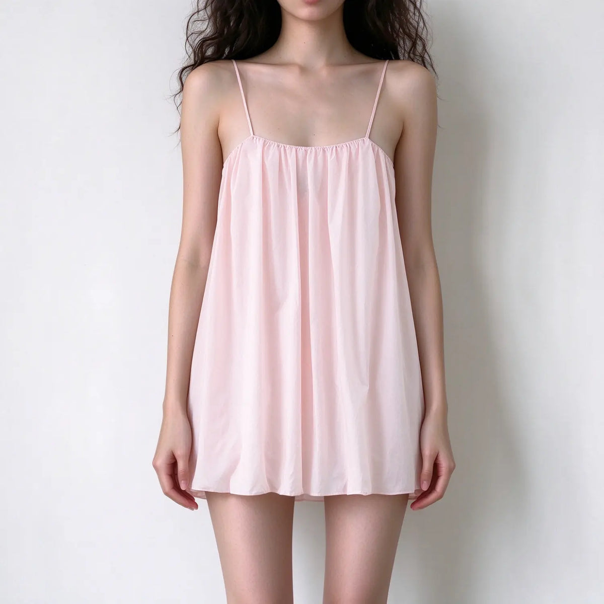 Model wearing light pink spaghetti strap mini dress, loose flowy a-line silhouette, gathered bust, simple casual sundress against plain white background