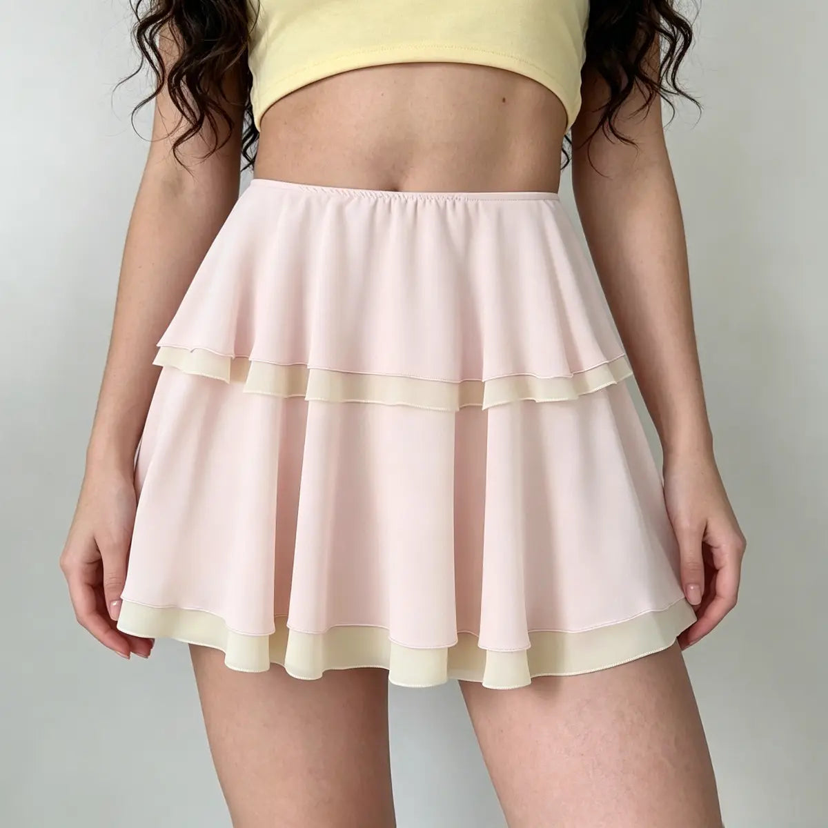 Model wearing a light pink tiered chiffon mini skirt with contrast beige trim, showcasing the flowy two-layer design and elasticated waistband.