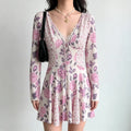 Model wearing a long-sleeve mini dress with a light pink base and large purple floral print, featuring a lace-trimmed V-neck and front tie detail.