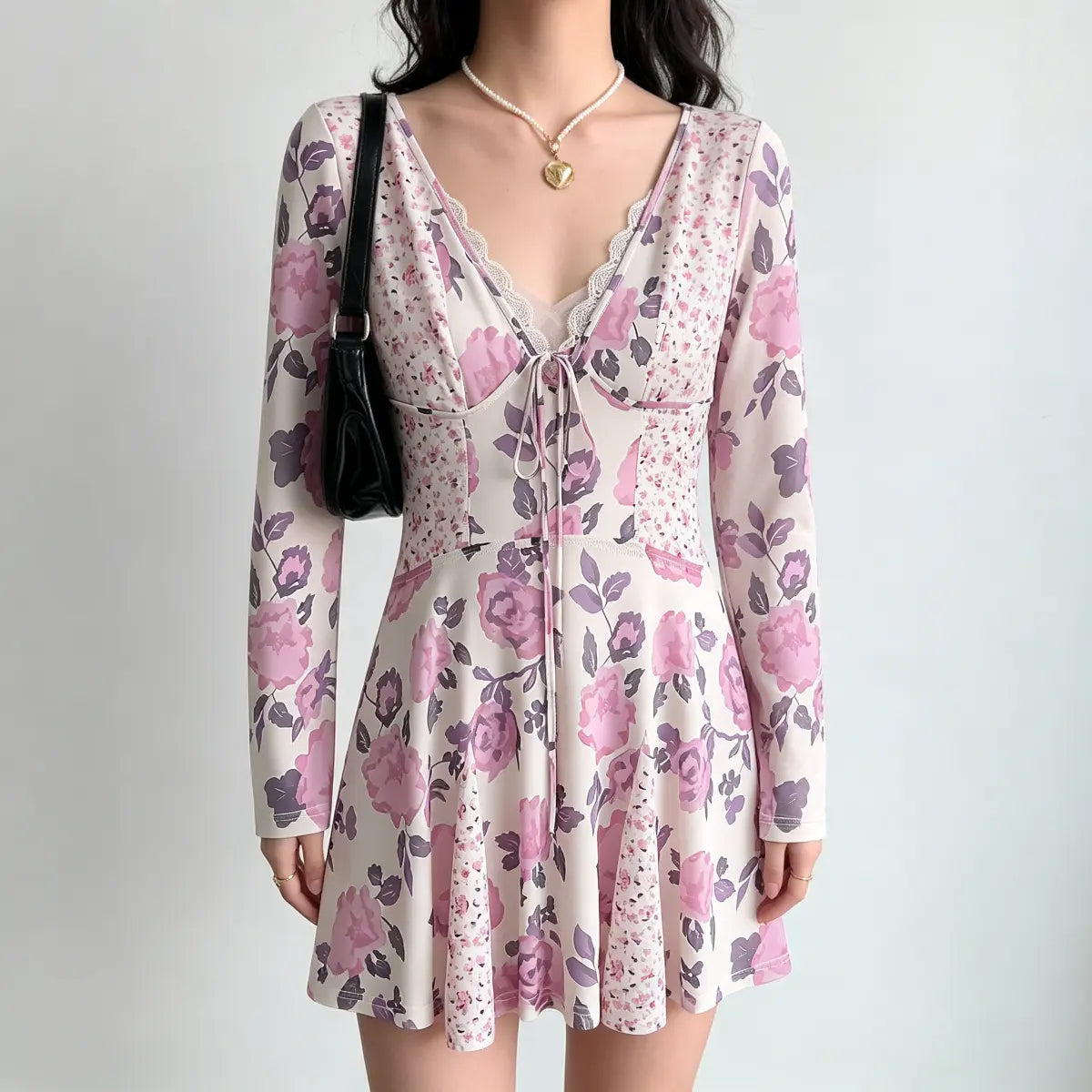Model wearing a long-sleeve mini dress with a light pink base and large purple floral print, featuring a lace-trimmed V-neck and front tie detail.