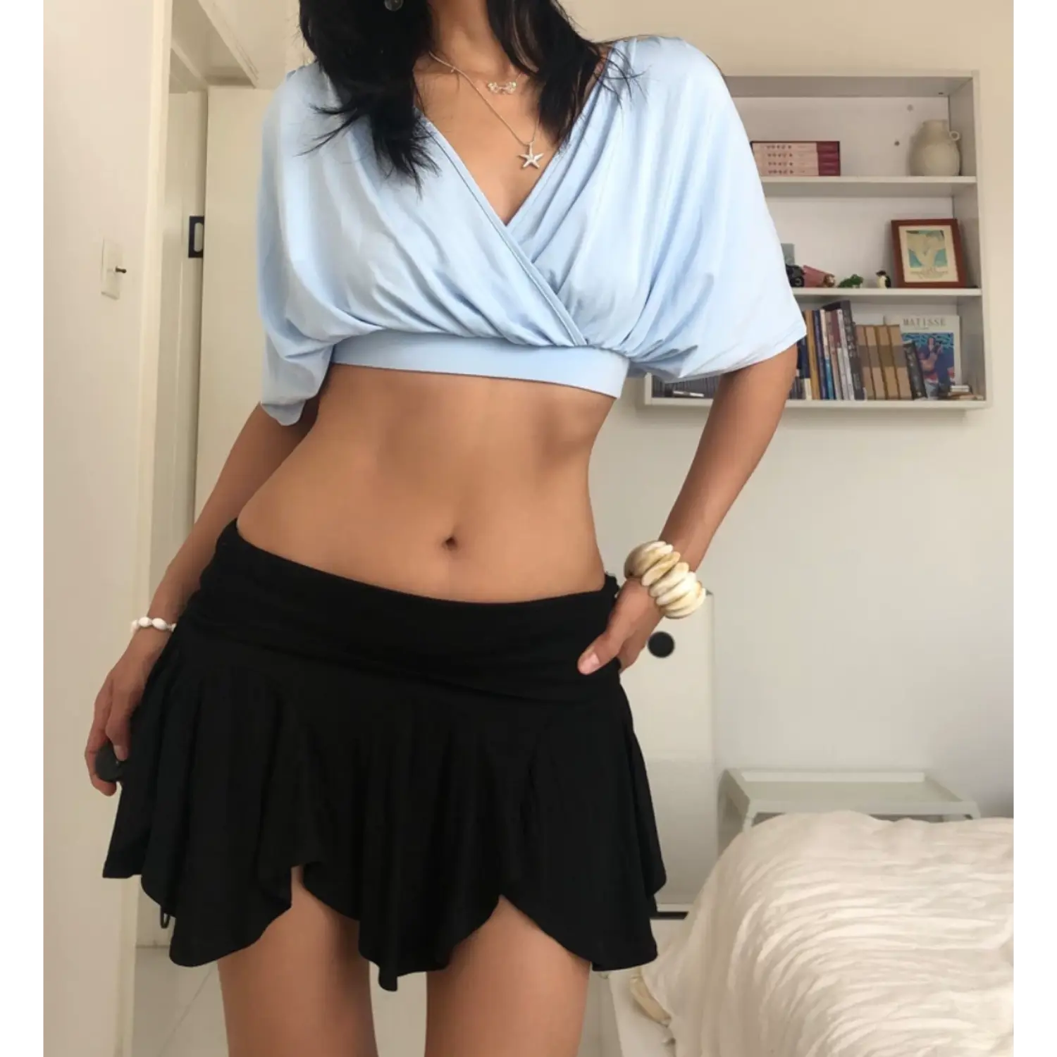 Medium full casual indoor lifestyle shot of sky blue wrap cropped blouse, model placing one hand lightly on hip, oversized flowy batwing sleeves, front surplice ruching for figure flattery, paired with black asymmetric ruffle mini skirt, shell bead bracelets, simple white room decor background