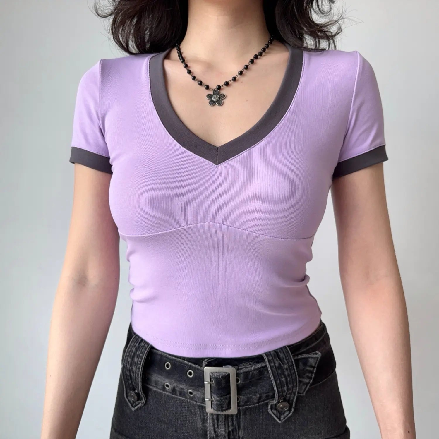 Lifestyle shot of a woman wearing a lilac fitted V-neck tee with dark gray trim, paired with dark denim jeans