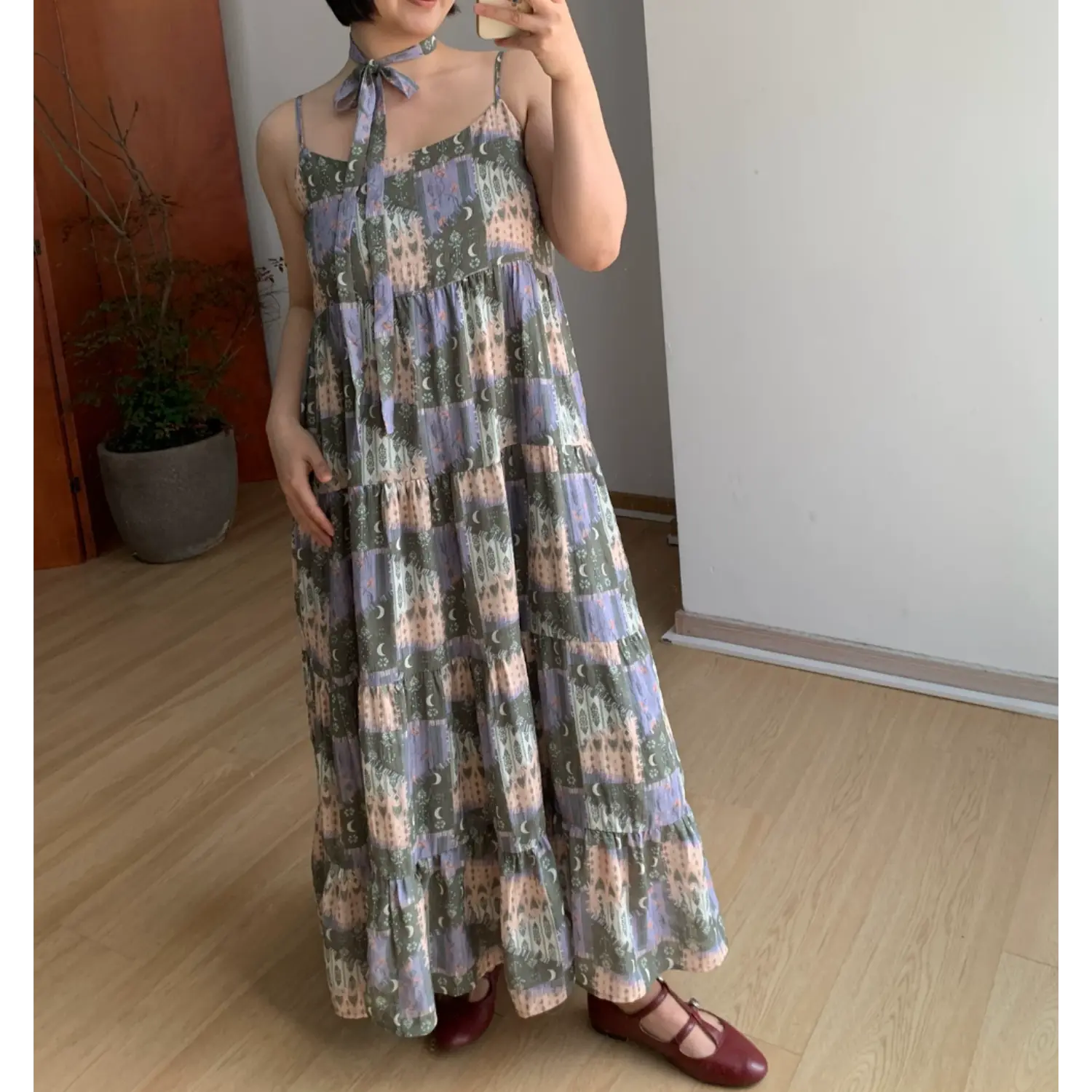Indoor mirror partial full shot of flowy tiered cami maxi dress with hidden side pocket (model resting one hand inside pocket), retro muted tone fantasy print, matching fabric neck choker, thin spaghetti straps, voluminous multi-layer ruffle floor skirt, burgundy mary jane flats, simple potted plant room decor