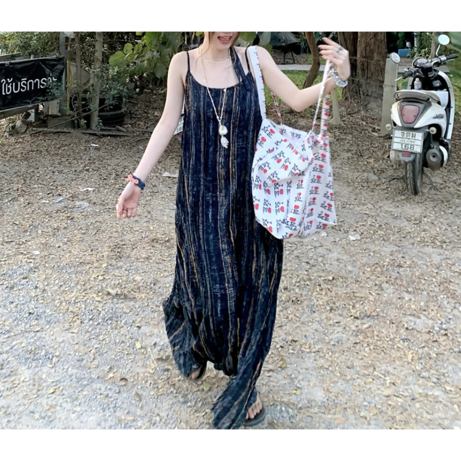 Walking outdoor lifestyle shot of sleeveless wrinkle texture maxi cami dress, vintage faded vertical stripe print on deep navy base, thin spaghetti straps, flowy floor-grazing skirt, model carrying white graphic canvas tote bag, tropical roadside greenery & white scooter background
