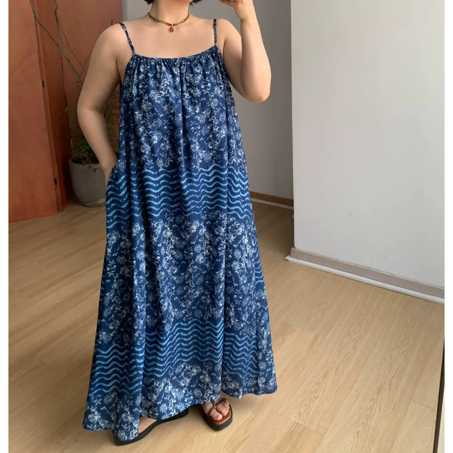Indoor mirror partial full shot of flowy batik slip maxi dress, model slipping one hand into hidden side pocket to show relaxed roomy silhouette, two-tier floral & wavy stripe batik print, thin spaghetti straps, lightweight wrinkle-resistant fabric, neutral minimalist room decor