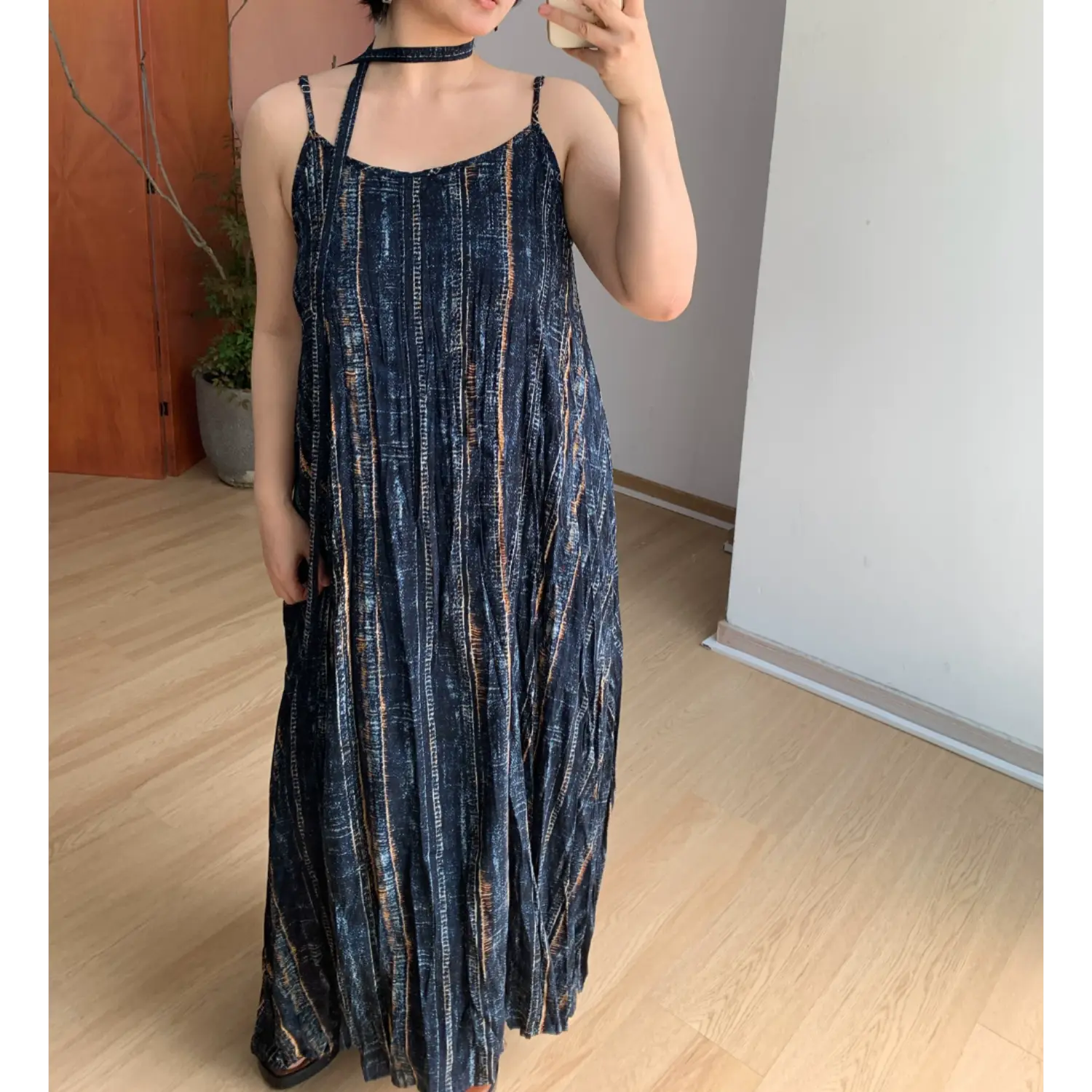 Front indoor mirror partial full shot of wrinkle textured striped cami maxi dress, hidden side pocket detail (model resting one hand inside pocket), matching fabric choker neck tie, thin spaghetti straps, flowy floor-length skirt, neutral minimalist home interior