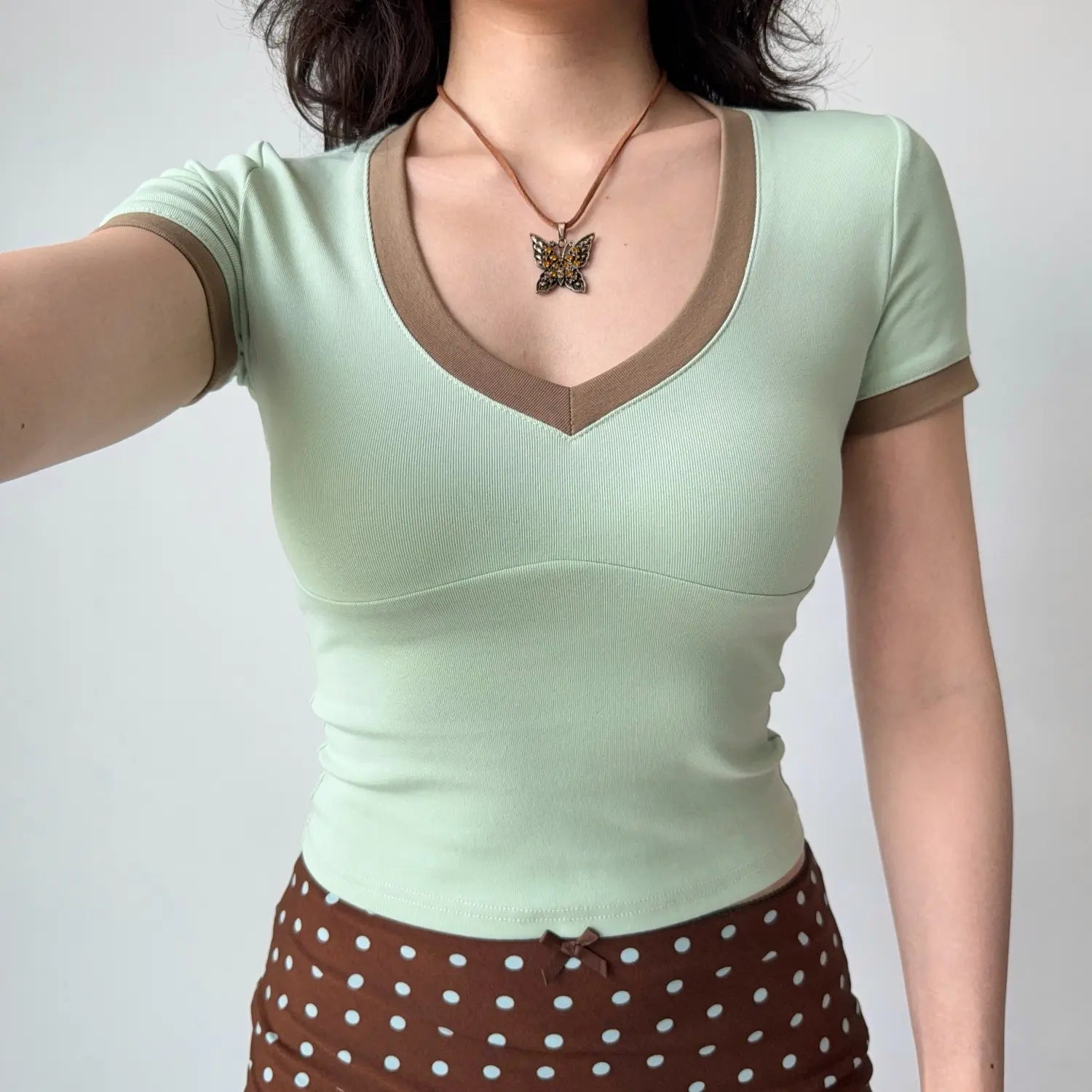 Selfie shot of a woman in a mint green fitted V-neck tee with brown trim, wearing a butterfly necklace