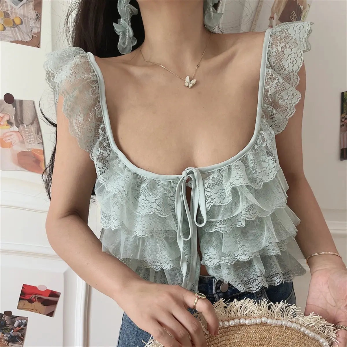 Close-up view of a woman wearing a mint green layered lace camisole with a front tie, paired with jeans and holding a straw hat.
