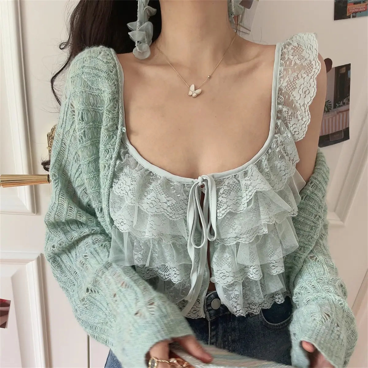 Angled view of a woman wearing a mint green layered lace camisole with a front tie, paired with a matching knit cardigan and jeans.
