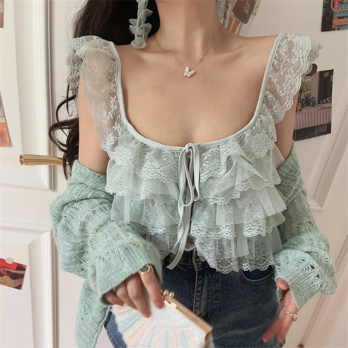 Front view of a woman wearing a mint green layered lace camisole with a front tie, paired with a matching knit cardigan, jeans, and holding a pastel clutch.
