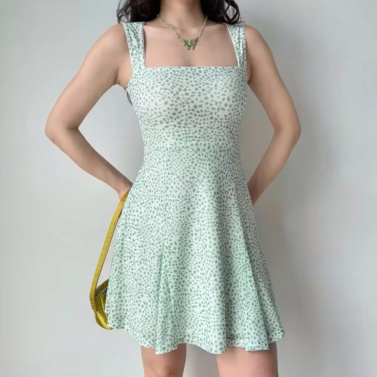 Woman wearing a mint green and white leopard print mini dress with a square neckline and fitted flare silhouette, styled with a yellow bag and butterfly necklace.