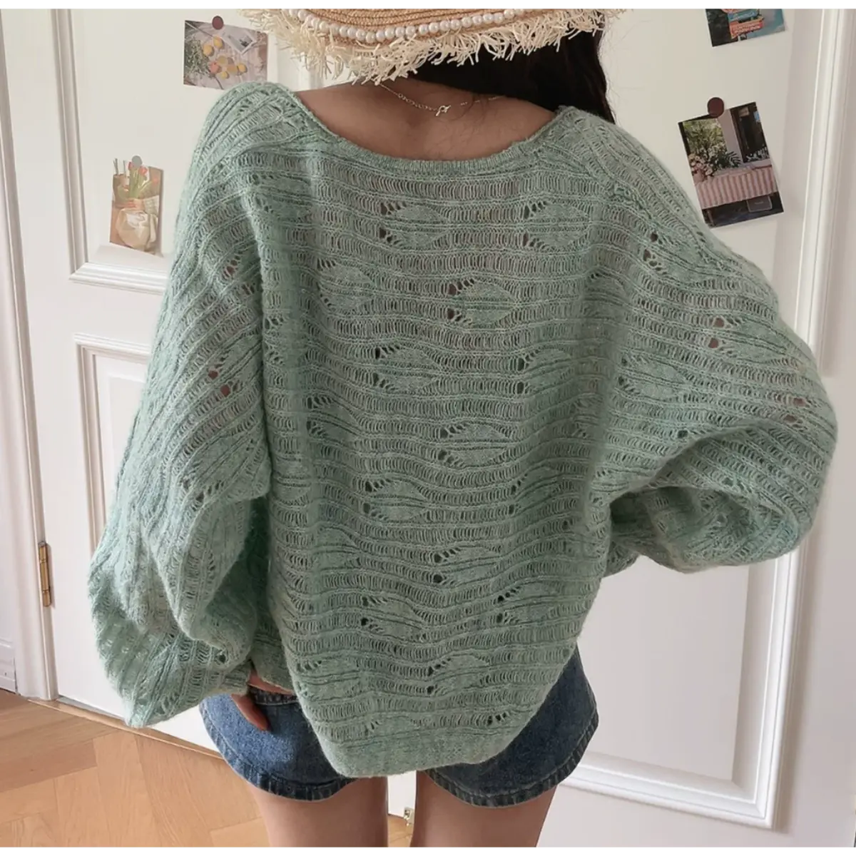 Back view of a mint green openwork knit cardigan showing its detailed knit pattern, worn over denim shorts and paired with a straw hat.