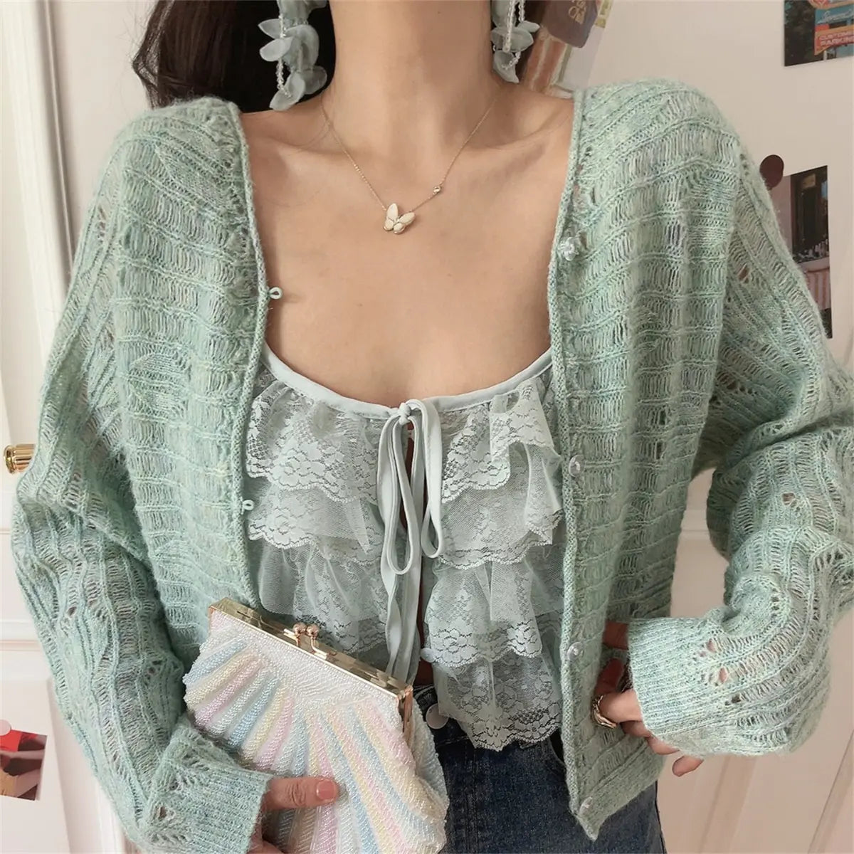 Angled view of a mint green openwork knit cardigan worn over a matching lace ruffled camisole, paired with jeans, holding a pastel rainbow beaded clutch.