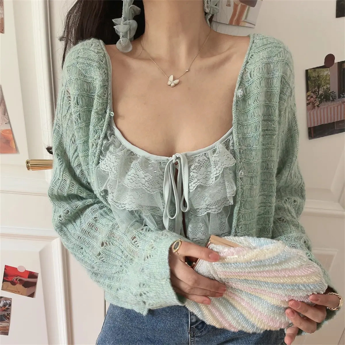 Close-up view of a mint green openwork knit cardigan worn over a matching lace ruffled camisole, paired with jeans, holding a pastel rainbow beaded clutch.