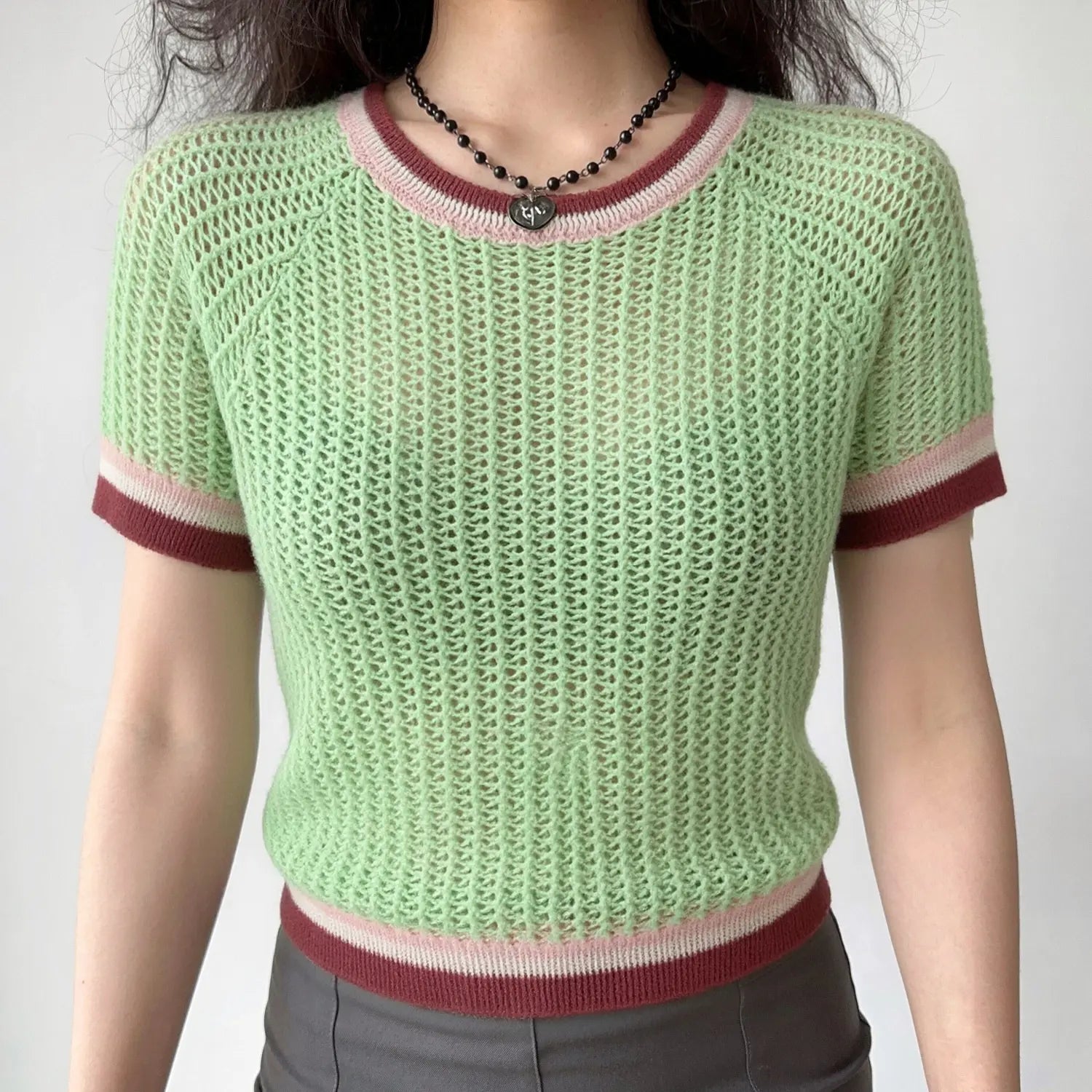 Front lifestyle shot on model, light mint green openwork mesh knit short sleeve top, round neck with pink-white-burgundy striped trim, raglan sleeves, cropped fitted hem, matched with grey bottoms and heart pendant necklace, casual summer daily outfit