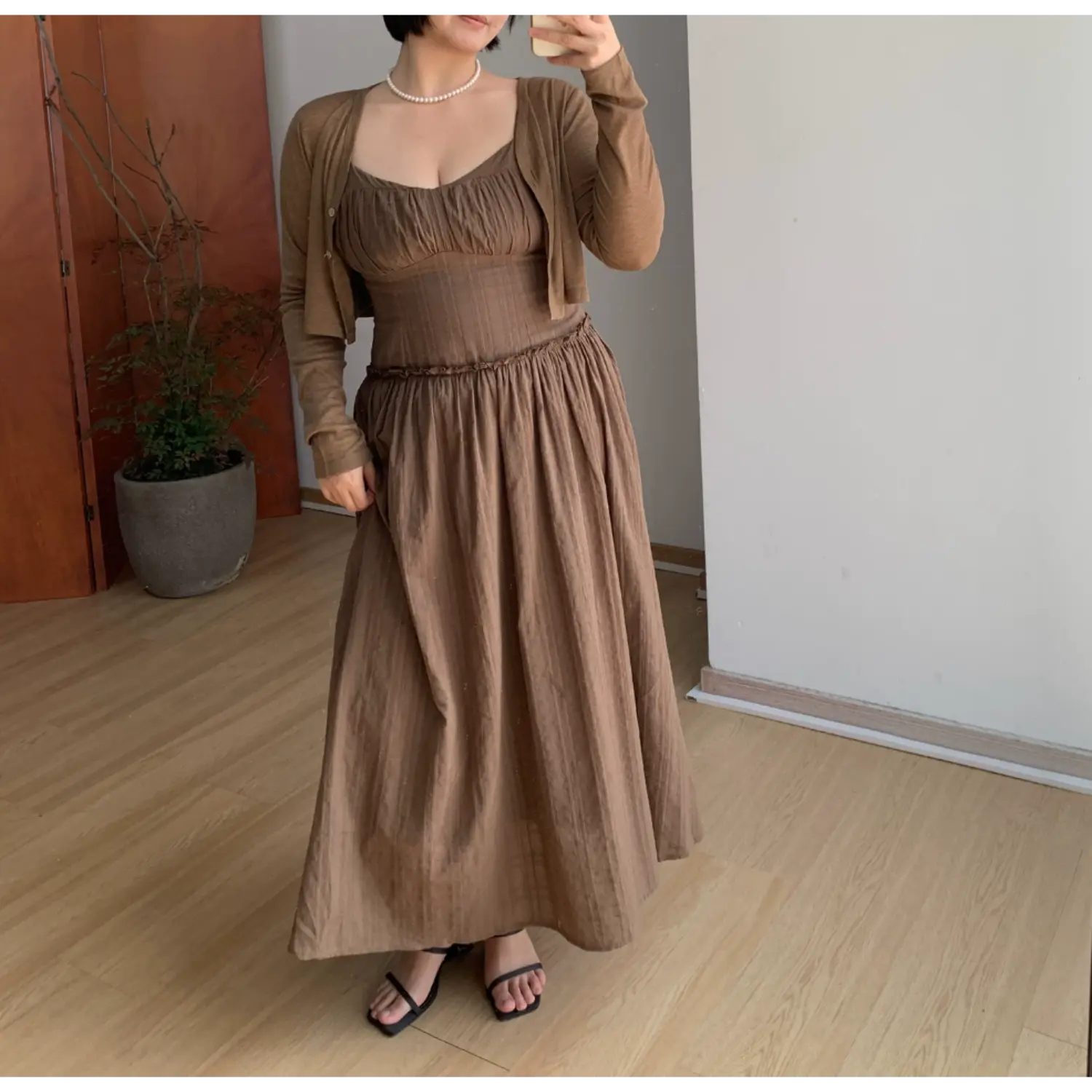 Indoor mirror partial full shot of brown textured cami maxi dress layered with matching cropped knit cardigan, model slipping one hand into hidden side pocket to show relaxed roomy skirt silhouette, ruched sweetheart bust, ruffle waist seam, black thin slide sandals, neutral minimalist home interior