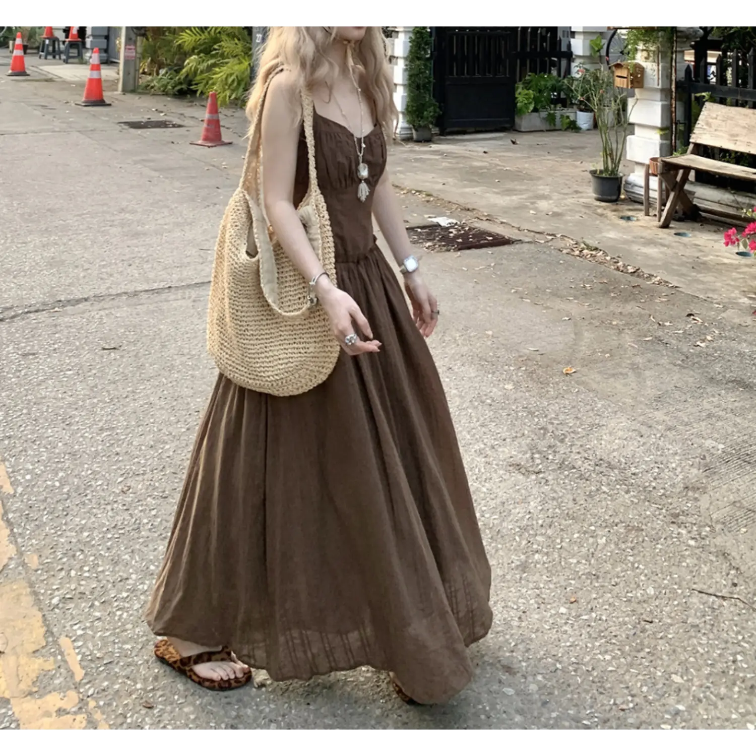 Side profile outdoor street full shot of textured empire waist cami maxi dress, soft matte brown fabric with faint vertical texture, gathered bust, loose swing floor skirt, large woven straw shoulder bag, leopard print thong slides, residential street traffic cones & green foliage scenery