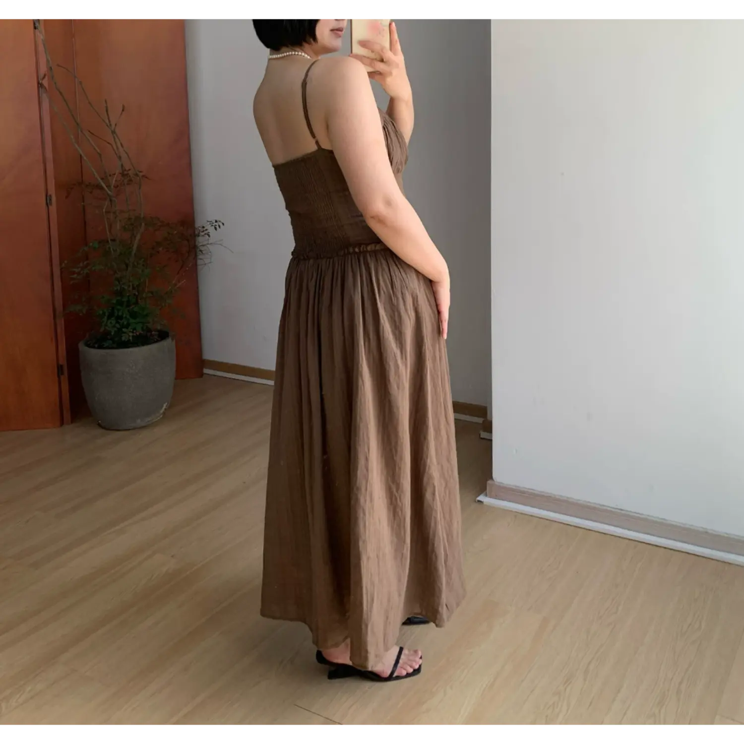 Side-back indoor mirror shot of textured brown empire waist maxi dress, stretch smocked back bodice for flexible fit, thin adjustable spaghetti straps, full billowy gathered long skirt, soft matte wrinkle-resistant fabric drape, black thin heel slide sandals