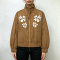 Female model wearing brown faux suede bomber jacket with white floral embroidery, front full view