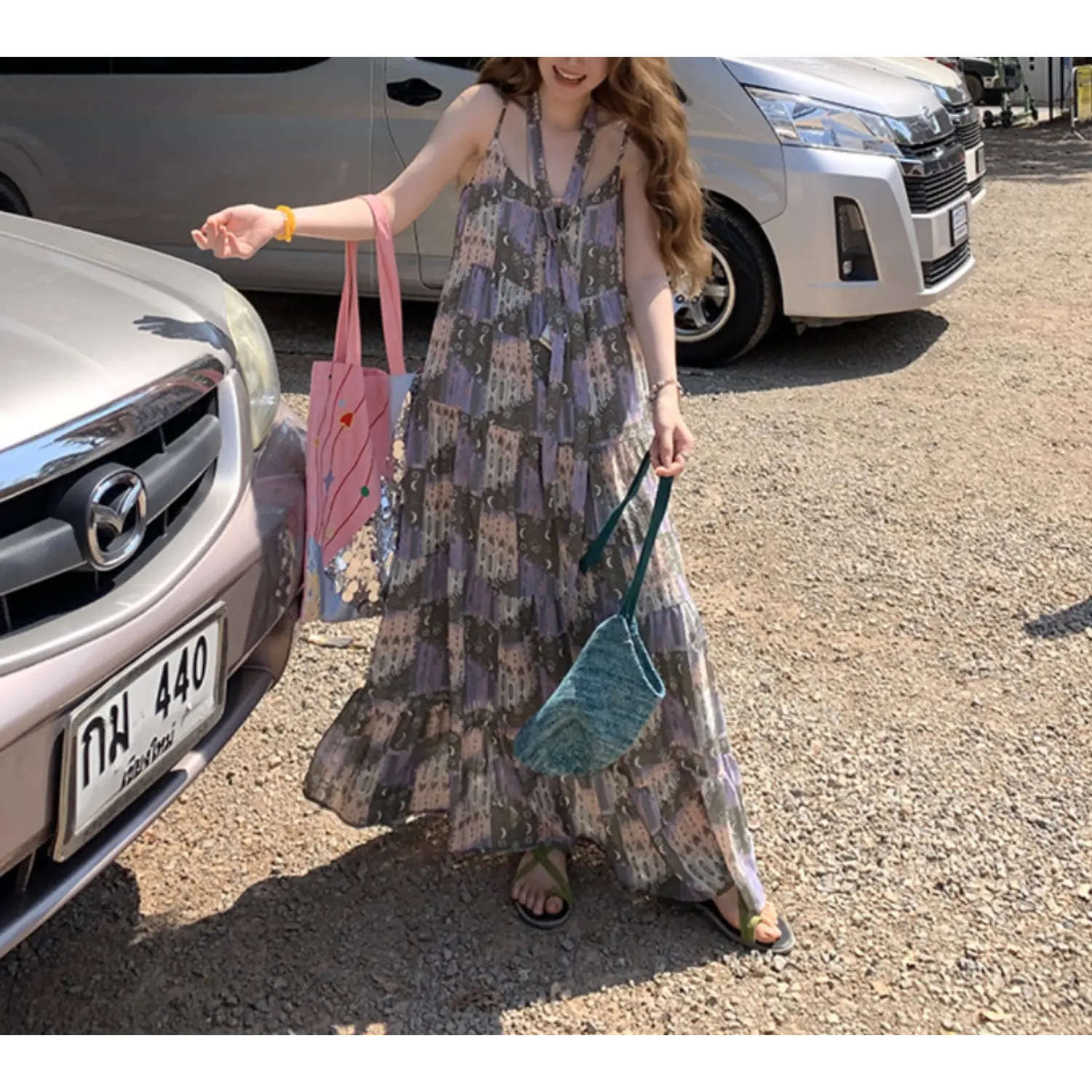 Static outdoor gravel parking lot full shot of loose tier cami maxi sundress, retro moon castle collage print, multi ruffle tier flowy floor skirt, thin adjustable cami straps, model carrying pink graphic tote & teal woven crossbody bag, green slide sandals, silver minivan background, bright natural daylight