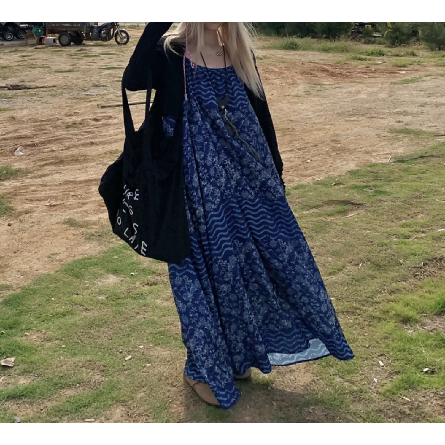 Three-quarter outdoor grass shot of oversized batik cami maxi dress with black sheer cardigan overlay, two-tone floral & wavy stripe print, loose wide swing silhouette, thin adjustable cami straps, black canvas shoulder bag, natural hazy daylight, dry grass & motorbike background