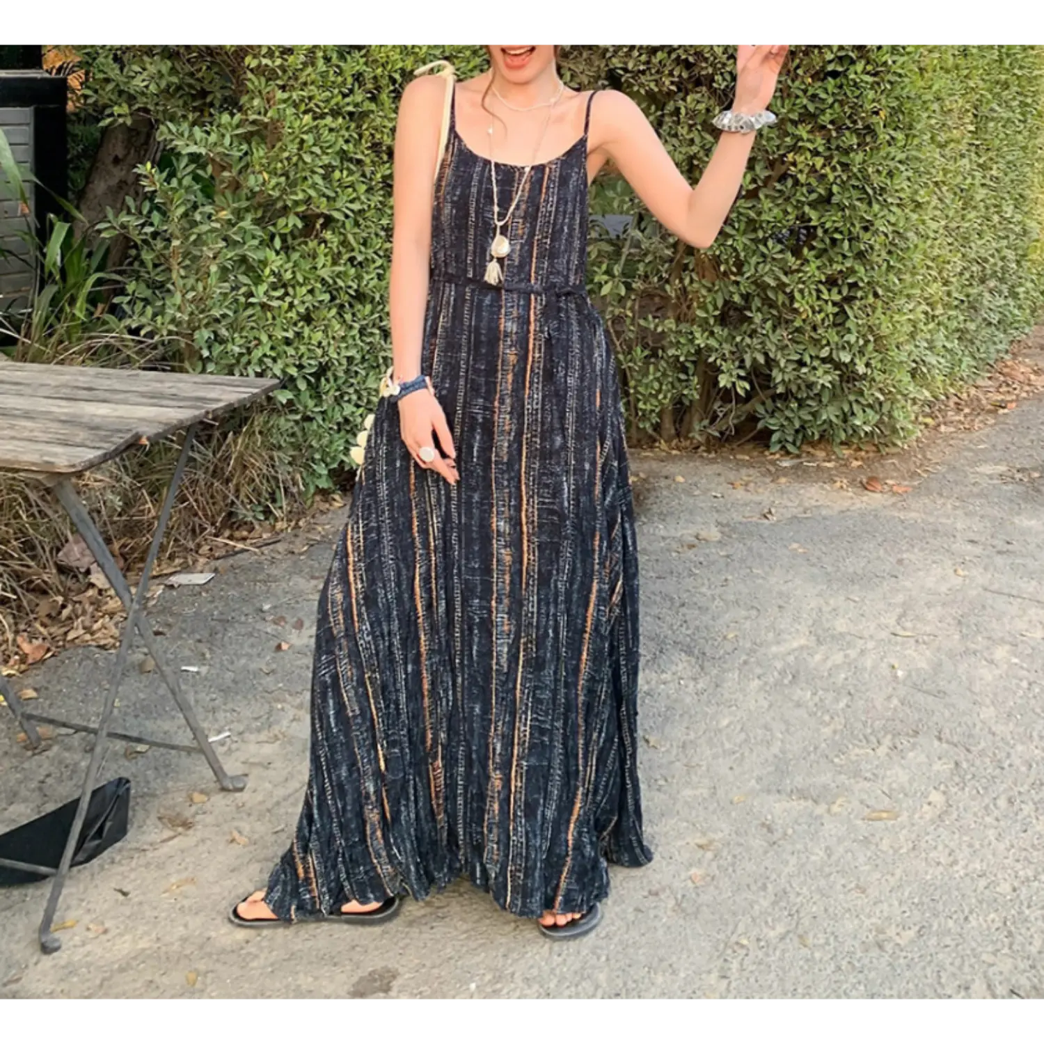 Static outdoor garden full shot of textured striped slip maxi dress, matching self-fabric waist tie to define loose silhouette, thin adjustable cami straps, soft crinkle wrinkle fabric, styled with black flat slide sandals, trimmed green hedge & wooden table backdrop