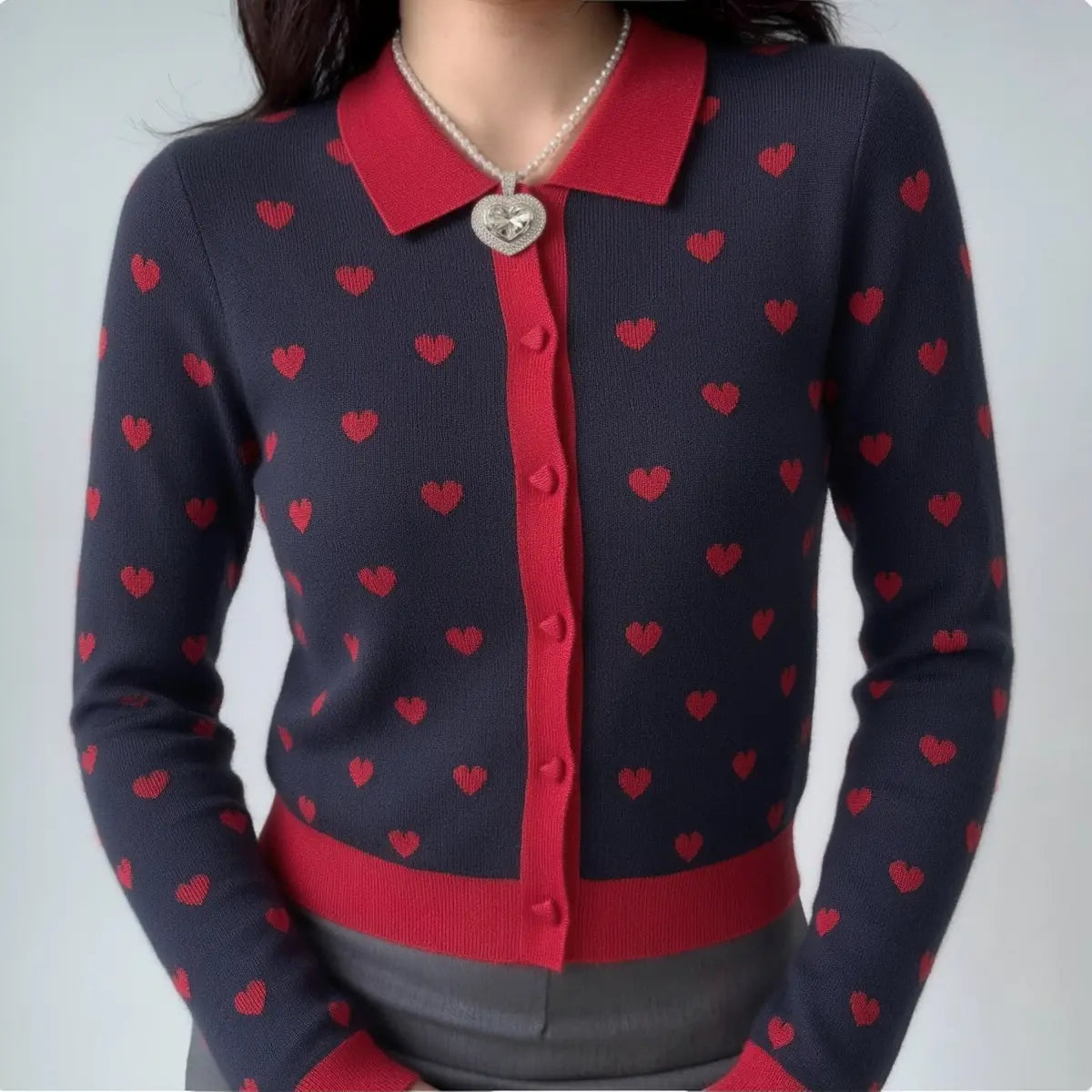 Full-body shot of a woman wearing a navy and red heart-patterned knit cardigan with a pearl necklace and gray bottoms.