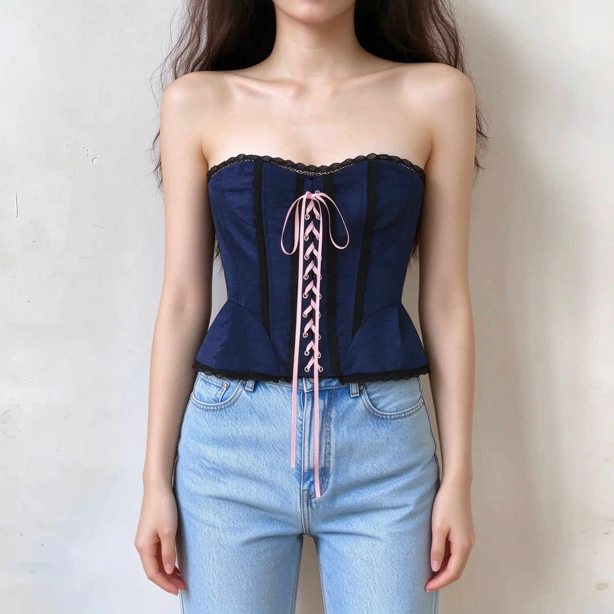 Model wearing navy strapless corset top, sweetheart neckline, black lace trim, front pink lace-up detail, peplum hem, fitted structured silhouette
