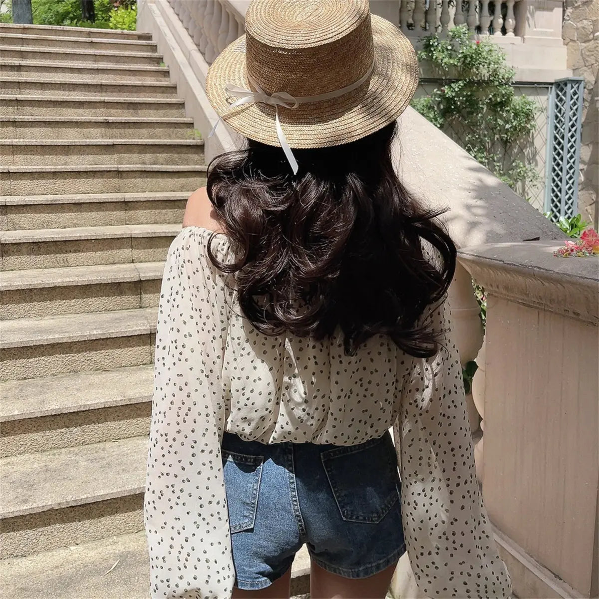 Back view of the off-shoulder cream blouse with black dot print, worn with denim shorts and a straw hat