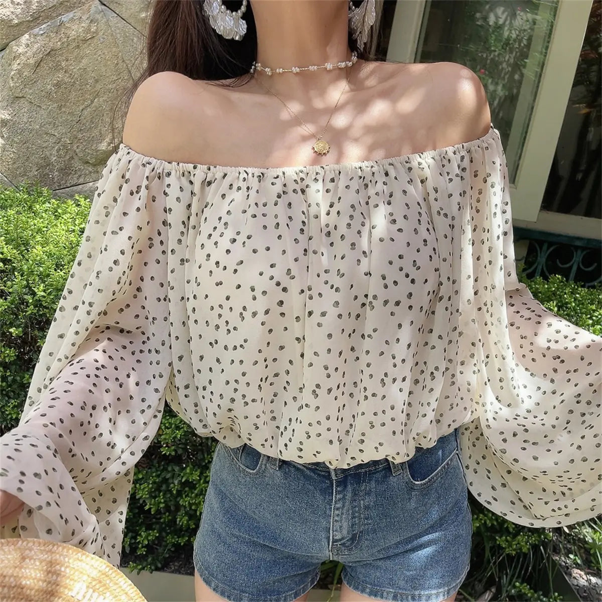 Close-up outdoor shot of the off-shoulder cream blouse with black dot print, worn with a sun necklace and floral earrings