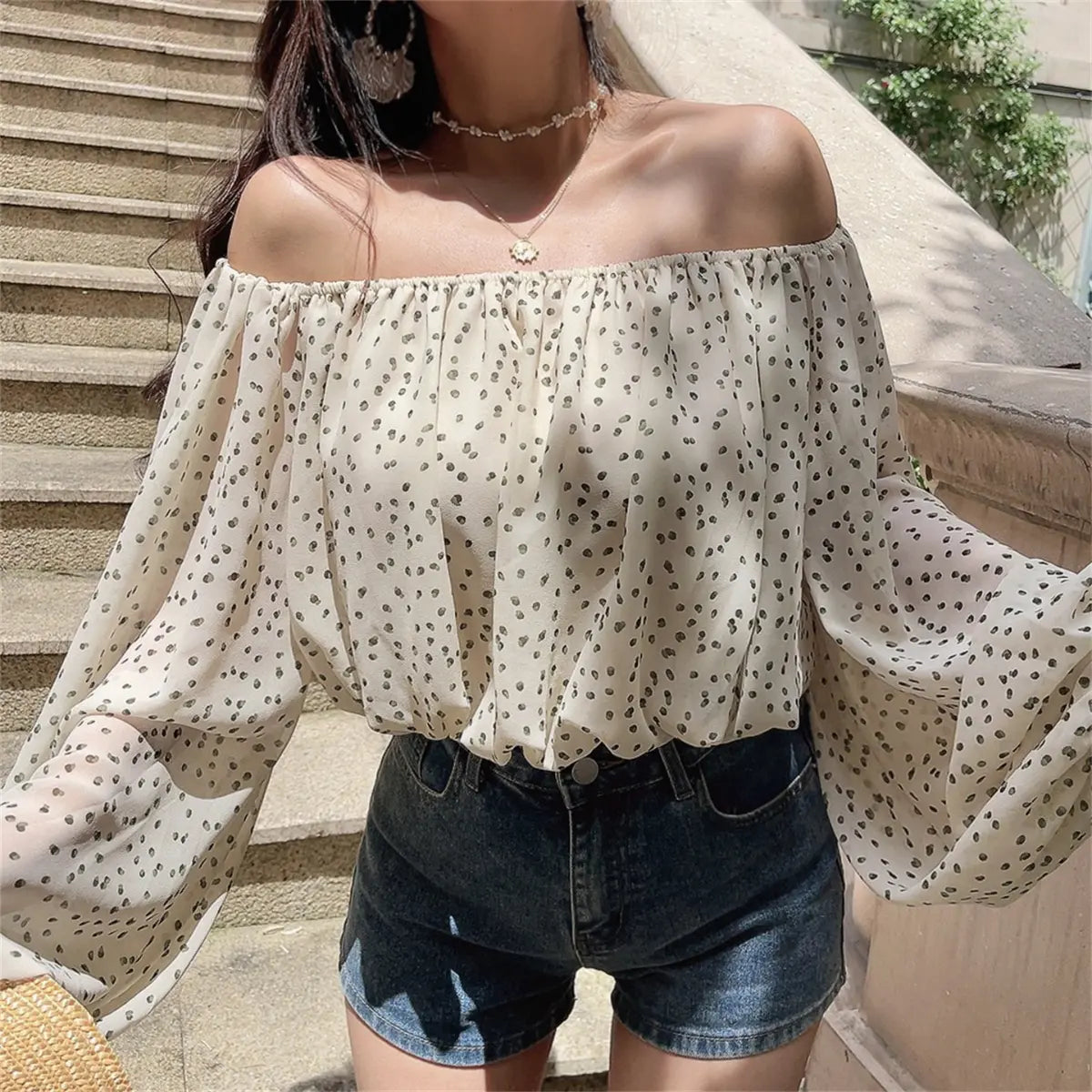 Outdoor shot of the off-shoulder cream blouse with black dot print, styled with denim shorts on stone stairs