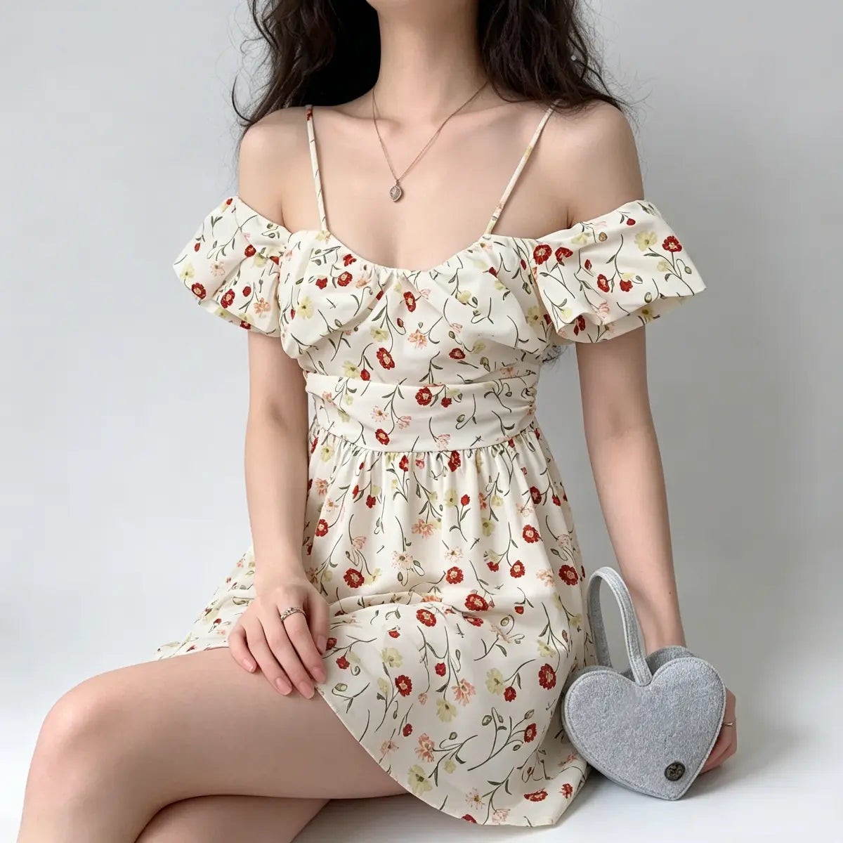 Seated view of a model wearing an off-shoulder floral mini dress, holding a heart-shaped bag beside her.