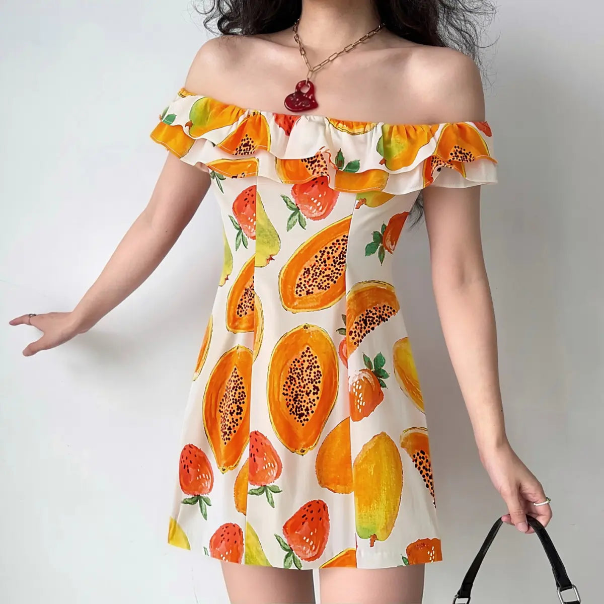 Model wearing off-shoulder fruit print mini dress, styled with a black bag for casual summer look.
