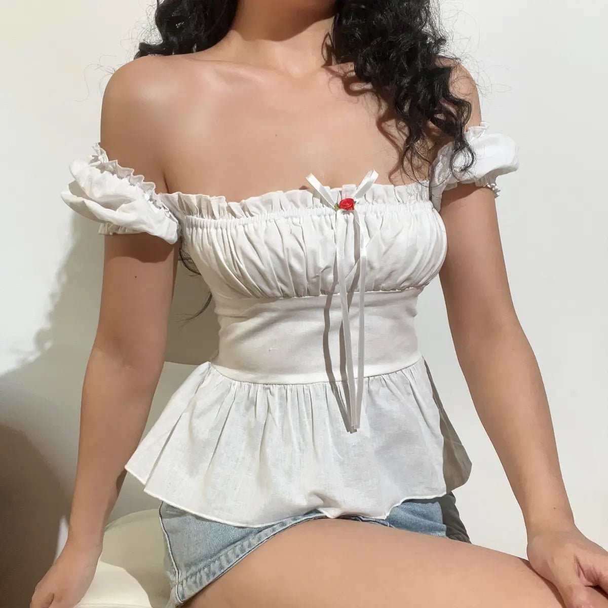  Close-up seated view of a white off-shoulder peplum top, emphasizing the ruffled sleeves and rose-accented ribbon detail.