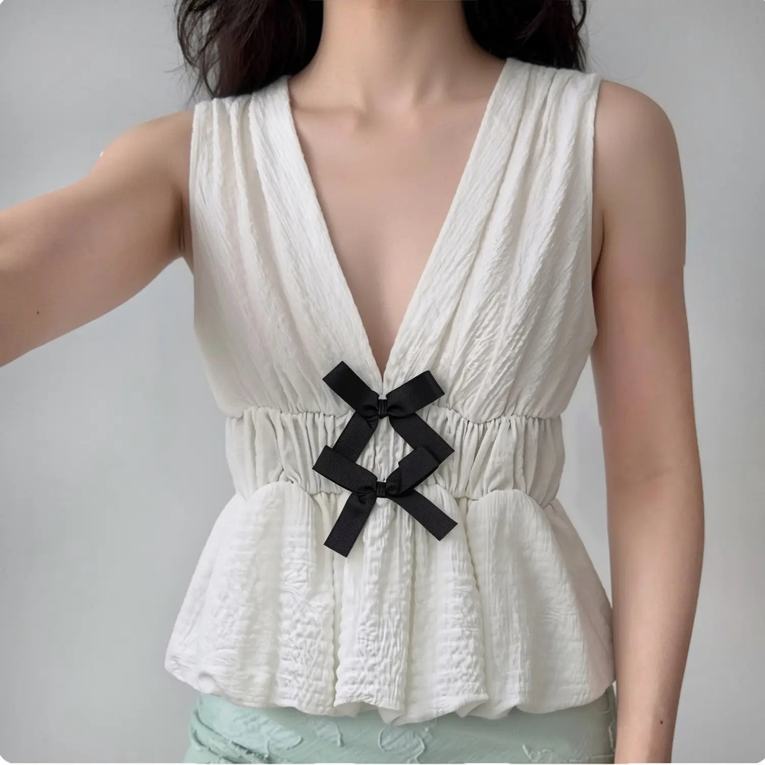 Lifestyle wearing shot of off-white crinkled fabric sleeveless peplum top with black bows, paired with light bottoms