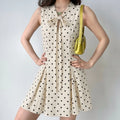 Front daily lifestyle shot of cream polka dot sleeveless mini dress, stand collar front self-fabric tie bow hollow detail, tailored fitted waist, swing flared skirt, matched with yellow shoulder bag, gentle vintage daily collocation